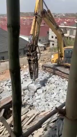 The video shows a yellow Komatsu excavator breaking rocks. The excavator has a hydraulic hammer attachment that is being used to break up a pile of large, gray rocks. The rocks are on the ground within a construction site, and there are bamboo scaffolding poles in the foreground. In the background, there are houses with red tile roofs.  The weather appears to be sunny. The excavator is stationary during the video and the hammer moves up and down repeatedly on the rocks.