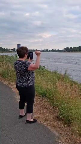 A person is standing on a paved path next to a body of water, facing away from the camera. They are wearing a black and white patterned short-sleeved shirt and black pants. Their right arm is raised, and they appear to be waving or signaling. The person is holding a smartphone in their left hand, possibly recording or taking a picture. In the background, a wide river flows, and on the opposite bank, there are trees and buildings, including a tall tower and a large passenger ship. The sky is cloudy. The overall lighting suggests it is late afternoon or early evening.