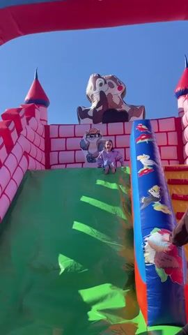 A young girl with long dark hair slides down an inflatable slide. The slide is part of an inflatable castle with images of cartoon chipmunks. The girl is wearing a purple shirt, jeans, and pink shoes. She is smiling as she goes down the green slide. An adult's arm is visible to the right, appearing to assist or guide the girl. The sky above is clear and blue.