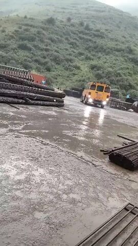 A yellow vehicle with flashing lights is moving on a wet, paved surface. The vehicle is facing away from the camera, and its brake lights are illuminated. The surface appears to be a storage or industrial area, with stacks of metal rods or pipes visible on the left and right sides of the frame. In the background, a steep, vegetated hillside rises under an overcast sky. The overall impression is of a functional, possibly industrial or construction site, with the vehicle moving in what appears to be a controlled manner.