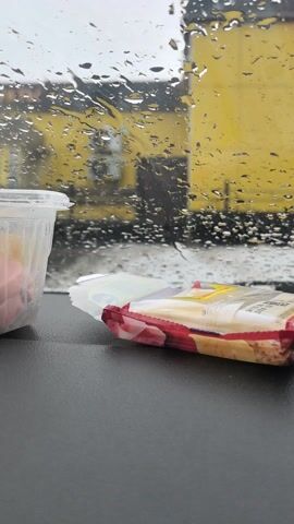 From inside a car, a view through a rain-streaked windshield shows a bright yellow building with a black structural element in the background. On the dashboard, there is a clear plastic cup and a red-and-white-packaged snack that is partially opened. The music is playing in the background.