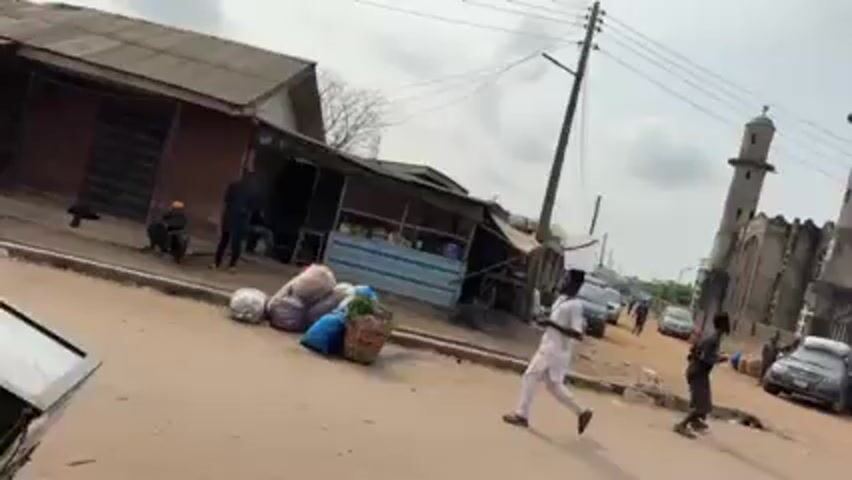 The video shows a street scene in what appears to be a village or town in Africa. In the background is a large, grey building with two tall towers, which appears to be a mosque. In the foreground, people are walking along the street. One person carries a bucket on their head, while another pushes a cart. There are several cars parked along the side of the street, and a few small shops are visible. The sky is overcast.