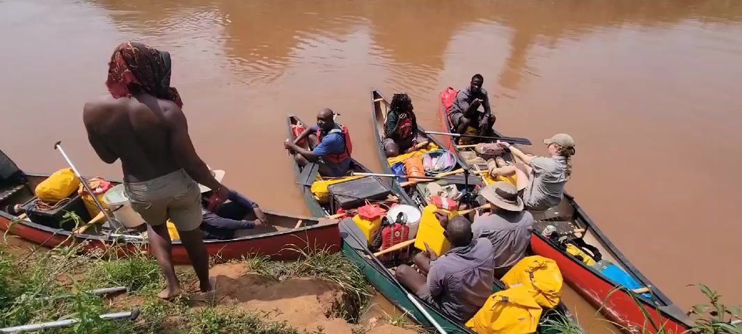 Several people are preparing to embark on a river journey in canoes. Three canoes are visible, each loaded with supplies and people. Two people are sitting in the first canoe, with one wearing a life jacket. In the second canoe, there are three people, one of whom is wearing a life jacket. The third canoe contains four people, one of whom is adjusting equipment. The river is a muddy brown color, and the shoreline is grassy. A shirtless person wearing shorts and a headscarf stands on the shore, apparently involved with the preparations. The surrounding environment includes trees and foliage.