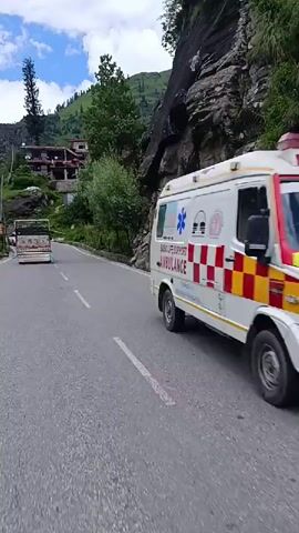 The video captures a scenic drive through a mountainous region. A paved road winds through lush green hills and trees. On the right side of the road, a rocky cliff face rises steeply, partially covered in vegetation. On the left, there are more trees and some buildings nestled on the hillside. A truck decorated with colorful paintings is traveling ahead on the road, and a motorcycle is visible behind it. The sky is partly cloudy with patches of blue. The overall impression is one of a beautiful and somewhat remote natural landscape.