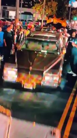 The video depicts the aftermath of a vehicle accident at night. An overturned car lies on the street with its undercarriage exposed, its wheels illuminated by lights. Several people are gathered around the vehicle, seemingly observing the scene. Streetlights and other vehicles are visible in the background. The ground is wet, possibly due to water or other fluids from the damaged car.