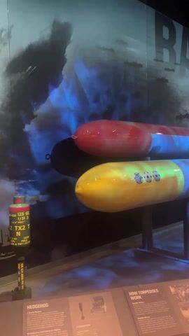 The video shows an exhibit on torpedoes and other naval armaments, possibly at a museum. There are two torpedoes on display, one painted red and another painted yellow and blue, resting on metal stands. Behind the torpedoes is a backdrop with the text "BATTLE OF THE A..." and a scene suggesting a naval battle. To the left of the torpedoes is a tall cylinder with text on it, and in front are information panels. To the right of the torpedoes, there are boxes labeled "ANCHOR MK 6". Further back, there's a black spherical object suspended from the ceiling and an anti-aircraft gun. The lighting in the exhibit is low, with spotlights highlighting the displays.