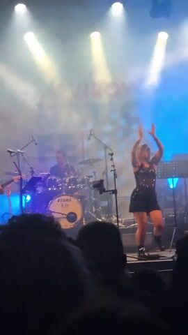 A live music performance is captured from the perspective of an audience member. The stage is lit with bright blue spotlights and smoke. In the center of the stage, a female performer in a black outfit with embellishments dances energetically with her arms raised. To her left, a drummer is seated behind a drum kit. To her right, a male guitarist plays an instrument. Other band members and equipment are visible on stage. The foreground is filled with the silhouettes of the audience, some with their hands raised. The video is very shaky.