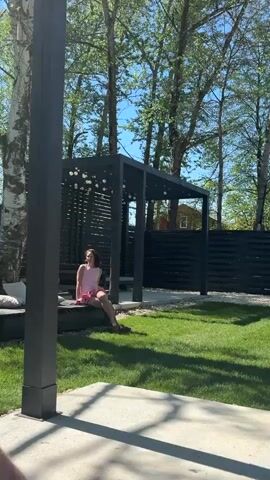 A wide shot reveals a backyard scene bathed in sunlight, featuring a fair-skinned, young woman wearing a pink dress and matching shoes. She is seated on what appears to be a dark-colored seat under a black pergola adorned with spherical lights. The pergola sits on a patch of green lawn, with a dark, horizontally-slatted fence behind it. Beyond the fence, glimpses of a house are visible among tall, slender trees with green leaves, set against a clear blue sky. The foreground of the shot includes a section of a concrete surface and a vertical black post that partially frames the view. The camera slowly pans to the right, offering a broader view of the backyard setting, including more of the trees and the distant house.