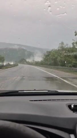 The video, filmed from inside a vehicle, shows a first-person perspective of driving on a wet, two-lane road. Raindrops are visible on the car's windshield, indicating current rainfall. The road ahead is asphalt, with clear white dividing lines and a solid white line on the right shoulder, bordered by a metal guardrail.

The surrounding landscape is hilly or mountainous, with dense green vegetation and trees lining the roadside. In the distance, hills are partially obscured by mist or low-hanging clouds, creating a subdued and somewhat hazy atmosphere. The sky appears overcast and gray.

The lower portion of the frame shows the car's dashboard and the top of the steering wheel, along with the base of the windshield wipers. No other vehicles or people are explicitly visible, suggesting a relatively quiet road. The time of day appears to be daytime, despite the cloudy weather. The scene is consistent with driving through a rural or mountainous area in Negushevo, Bulgaria, during a rainy period. There is no visible text in the video.