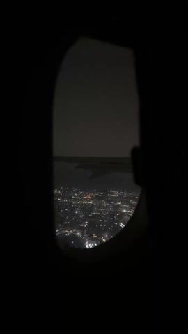From inside a darkened airplane cabin, the view through the window reveals a city at night. The city lights twinkle beneath a layer of cloud cover. A portion of the airplane wing is visible. The image is captured in dim lighting, emphasizing the city's glow.