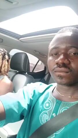 Two men are in a car. The driver is a young man with blonde dreadlocks and is wearing a grey tank top. He is singing along to music. The passenger, who appears to be recording, is wearing a turquoise traditional shirt and a seatbelt. The car has a sunroof.