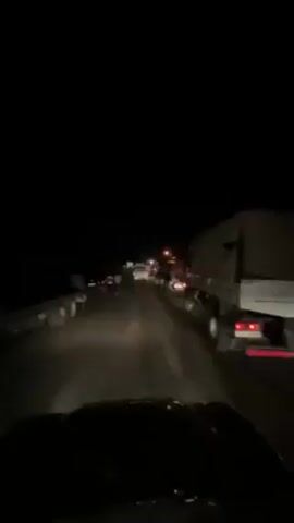 Here is a forensic description of the scene in the video.

The video shows a dark roadway with cars and trucks lining both sides, some with flashing emergency lights, indicating a blockage or accident. Several people are walking on the road, some seemingly in uniform, while others are dressed in casual clothing. The footage appears to be shot from inside a vehicle, capturing the view through the windshield as it slowly moves along the road. The overall atmosphere suggests a potentially serious event or emergency situation.