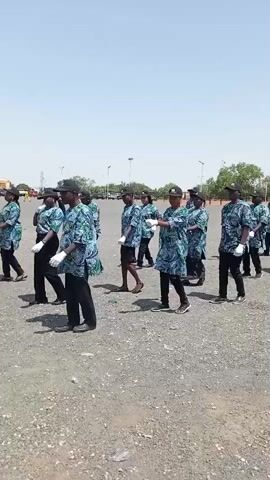 A group of approximately fifteen individuals, primarily men, are seen marching in formation across an open, unpaved area, likely a large gravel or dirt lot, under a clear, bright sky. They are dressed in matching short-sleeved shirts with a blue, green, and white patterned design, black trousers, black baseball caps, and white gloves. Their arms are swinging in unison as they walk forward in rows, suggesting a parade, ceremonial march, or drill practice. In the background, streetlights and distant trees are visible. The setting is outdoors and appears sunny and bright, indicating daytime. The scene takes place in Jimeta, Nigeria.