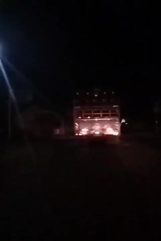 A truck with lights on top is visible from the rear at night. The truck appears to be moving. There are lights on the rear of the truck that are illuminated. The background is dark and appears to be a road.