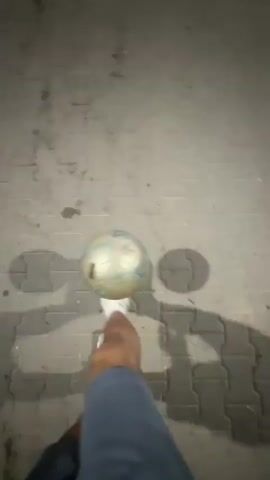 A first-person perspective video shows someone's left leg and foot wearing a white athletic shoe. The foot is positioned above a soccer ball on a paved ground with interlocking bricks. The person is likely performing a soccer drill or practicing juggling as the ball is manipulated by the foot. The lighting suggests it is evening or nighttime, with shadows cast by the person and the ball. The camera briefly blurs and then returns to showing the soccer ball rolling away on the ground, with the person's foot appearing near the ball.