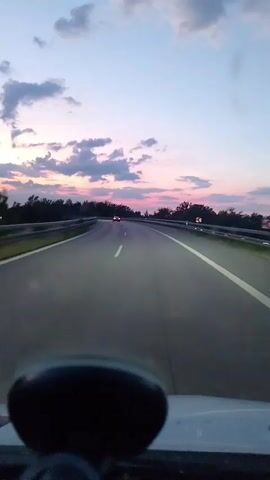 The video shows a view from the perspective of someone driving on a highway at dusk. The sky is a soft blend of blue and pink hues, with scattered clouds. Trees line the sides of the highway, and a guardrail is visible. In the distance, another vehicle's taillights are visible on the road ahead. The dashboard of the car, including a black object, is partially in the foreground. The video appears to be a static shot, with no visible movement apart from the implied forward motion of the vehicle.