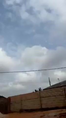 The video shows a yard with plants, a red car, and a person on a motorcycle. The yard has red soil and a concrete wall. There is a cloudy sky visible. The camera focuses on different plants in the yard, including a small pine tree. A child's voice can be heard speaking in the background.