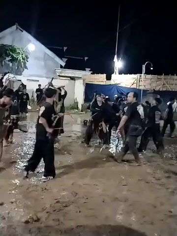 The video captures an outdoor night scene in Jenggrik, Indonesia, where a group of people are participating in a cultural or traditional event. The ground is extremely muddy, and most participants are visibly covered in mud. Many individuals, predominantly men, are wearing black t-shirts with a distinct golden emblem on the back, while others wear traditional Indonesian attire including long pants and sashes. Some are actively performing or dancing with energetic movements in the mud. One man in a light-colored top and darker pants is heavily splattered with mud, running through the scene. In the background, residential buildings and wooden structures are visible, illuminated by artificial lights, suggesting a community gathering. Another individual is seen holding a smartphone, presumably recording the event. The atmosphere appears lively and engaging, with the muddy conditions being a central element of the activity. The specific text on the golden emblem is not discernible.