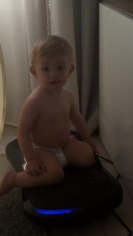 A baby is sitting on a black vibrating platform. The baby is wearing a diaper and is naked from the waist up. The baby has blonde hair and blue eyes and is looking down. The platform has colored lights that change from purple to red and yellow. The background shows a curtain and a white surface.