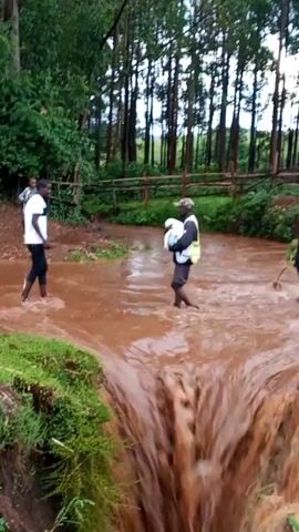 Two men are wading through a flooded stream. The stream has brown, muddy water, and a small waterfall is on the right.  One man wears a white t-shirt and black pants, and the other wears a dark shirt, shorts, a yellow safety vest, and a baseball cap. The second man is carrying something white on his back.  A child stands on the bank watching.  The area is surrounded by green grass and trees.  A split-rail fence is visible in the background.
