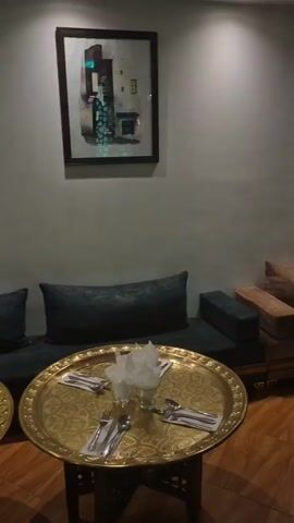 The video showcases the interior of a Moroccan-themed lounge. The room features low-set sofas upholstered in varying shades of brown and green, arranged around a series of ornately decorated brass tables laid out with silverware and napkins. The decor is detailed, including traditional Moroccan lamps and recessed wall niches displaying figurines and artifacts. The room appears to be set up for dining or lounging.