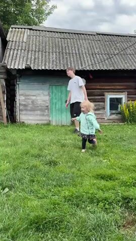Two children are playing with a blue ball in a grassy yard in front of two rustic wooden buildings. The building on the left has a red corrugated metal roof and is partially open on the left side, revealing what looks like a white appliance inside. The building on the right has a grey, textured roof and a green door. A large, shiny metal pot sits in the foreground to the left.

The taller child, a boy, is wearing a white t-shirt and dark shorts. He is in the process of kicking or throwing the blue ball, which is in the air above his head. The younger child, a girl, is wearing a light blue jacket, dark leggings, and pink shoes. She is running towards the ball, her arms outstretched. She appears excited.

The scene is outdoors, on a sunny day with scattered white clouds in a blue sky. The overall impression is one of playful activity in a rural setting. There is no visible text in the media file.