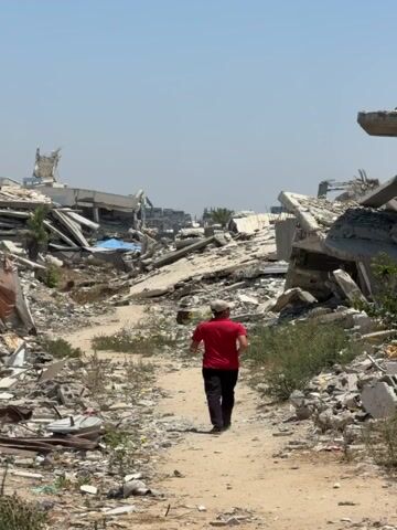 A person wearing a red shirt and a hat walks down a dirt path through an area devastated by destruction. The landscape is filled with rubble and debris from collapsed buildings. The sky is clear and blue, indicating a sunny day. The person is walking away from the camera.