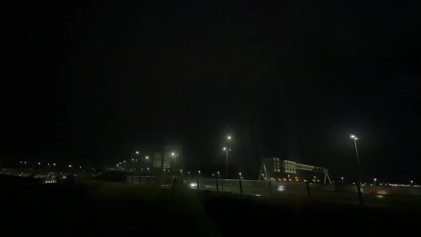 The video shows a nighttime scene, likely from the perspective of a moving vehicle. The sky is dark, with faint lights visible in the distance. Several streetlights illuminate a road or highway, casting pools of light. In the background, a large, illuminated structure, possibly a bridge or industrial building, is visible. The lights from the structure and the streetlights suggest an urban or suburban environment. There are some indications of vehicles on the road, with faint headlights and taillights. The overall impression is one of a low-light, possibly damp environment, with artificial illumination dominating the scene.