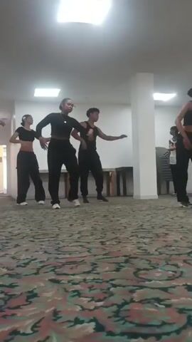 Four young people, two females and two males, are dancing in a room with a patterned carpet. They are all wearing black athletic wear, including crop tops and loose pants. The room is lit by overhead fluorescent lights and appears to be an indoor space with a white wall and a wooden table in the background. The dancers are performing synchronized movements, with some individuals moving their arms and torsos in unison. The camera is positioned low to the ground, giving a slightly distorted perspective of the scene.