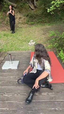 The video begins with a view of two women outdoors. One stands on a grassy slope, taking a photograph with a small electronic device. She is wearing a long black dress and black boots. The other woman sits cross-legged on a red pad on a wooden deck. She is wearing black pants, black boots, and a patterned jacket over a white top. The camera pans across the deck, revealing a group of people chatting and relaxing on chairs and lounging on the deck. A man walks by carrying a guitar case. The deck extends over a still body of water, the reflection of the trees on the far bank mirrored in the water. The video ends with a close-up shot of the still water and tree reflections.