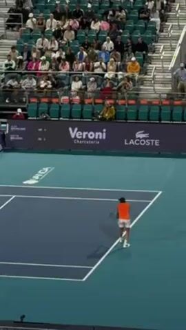 The video shows a tennis match in progress. A tennis player in an orange shirt and white shorts is preparing to serve the ball on a blue tennis court with white lines. Behind the player, there is a crowd of spectators sitting in tiered seating. A large advertising banner is visible in the background with the words "Veroni Charcuterie" and the Lacoste logo. The camera angle is from a high vantage point, providing a wide view of the court and surrounding area.