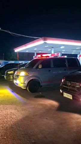 Two cars are parked at a gas station at night. The car in the foreground is a dark colored SUV. The car behind it is a silver minivan with red and orange stripes. The minivan has red flashing lights on its roof. The gas station has a red and white awning with lights. The sky is dark and there are some wires visible overhead. The ground is a dirt or gravel surface.