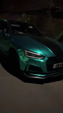 The video features several short clips of a custom-colored Audi S5 coupe parked outdoors at nighttime. The car is finished in an iridescent metallic green or teal, with a black stripe running down the center of the hood and a black roof. It is equipped with black multi-spoke wheels, a front splitter, and custom side skirts.

The car's LED headlights and taillights are illuminated, casting reflections on its glossy surface. It is parked alongside a brick wall and some dark foliage, indicating an urban or suburban setting.

The video presents different angles of the vehicle. The front view shows the Audi rings logo and the license plate "C9 YXC" (partially visible in the first clip). A side view reveals a "V6T" badge on the fender. The rear view clearly displays the "S5" badge, the Audi rings logo, the full license plate "C9 YXC", and quad exhaust tips. The final clips offer additional perspectives of the car's side profile.

No people are present in the video, and the car remains stationary throughout. The footage appears to be a showcase of the vehicle's unique aesthetic under low-light conditions. The location context for the media file is South Kesteven, United Kingdom.