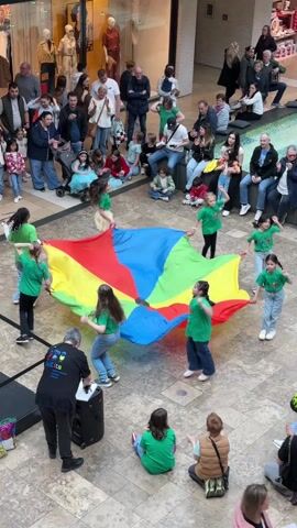 The video depicts an indoor scene, likely a shopping mall in Rheine, Germany, during the daytime. A group of approximately ten children, mostly wearing green t-shirts, are actively engaged in a game involving a large, colorful parachute. The parachute features distinct segments of red, blue, green, and yellow. The children hold the edges of the parachute, moving it up and down.

An adult male, wearing a black t-shirt with "Kids" partially visible on it, is positioned near the children, operating a black sound system consisting of a speaker and a control board. Other adults and children are observing the activity, some standing and others seated on benches in the background. The background features display windows of shops, showcasing mannequins and clothing, indicating a retail environment. The flooring is a light-colored tiled surface. The overall atmosphere is lively and joyful, with the children appearing happy and engaged in the parachute play, while the onlookers enjoy the spectacle. "Mid" is partially visible on a sign in the background.