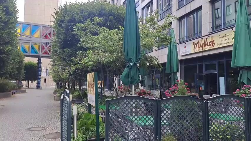 The video shows a street scene with a restaurant called Mythos on the right side. The restaurant has outdoor seating with green umbrellas and tables. There are also trees and plants in front of the restaurant. In the background, there is a modern building with a colorful glass structure. The street is paved, and there are some parked cars. The weather appears to be overcast.