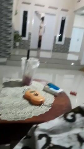 The video shows a close-up of a wooden table with various items on it, set against the backdrop of a house entrance. On the table, there's a plastic cup with a straw and a purple liquid, a small orange digital scale, a pack of cigarettes, and a blue lighter. A red object, possibly another cigarette pack, is visible further to the right. A portion of a patterned cloth is in the foreground, obscuring the lower part of the frame.

The house entrance in the background features a wide staircase leading up to a double white door, flanked by tall, narrow windows. The architecture suggests a residential setting, possibly in the evening or at night, given the artificial lighting. The overall mood is casual and relaxed, with the items on the table implying a moment of pause or a break. There are no visible text elements in the video.