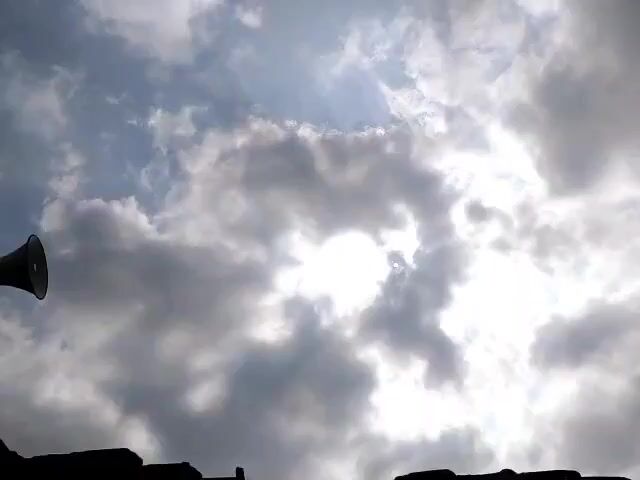 The video shows a low-angle shot of a cloudy sky with the sun partially visible behind the clouds. In the foreground, on the left side of the frame, a dark object is visible. It appears to be a megaphone or speaker mounted on top of a structure, possibly a building, which is silhouetted against the sky. The rest of the scene is dominated by the dynamic cloud formations and the bright sunlight breaking through them. There are no people visible in the video. The environment appears to be outdoors.