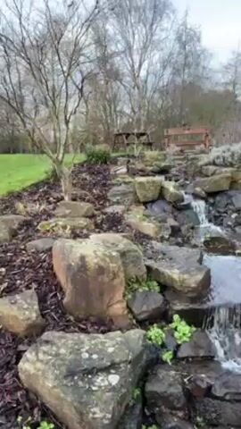 The video shows a small man-made waterfall cascading over rocks in a landscaped garden. The camera pans up to a wooden bridge with black railings. The bridge crosses the stream and leads to a paved path through the garden. There are trees, green grass, and buildings in the background. The sky is overcast.