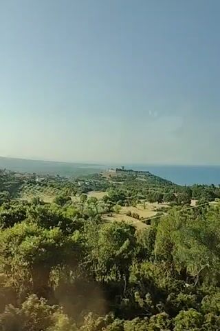 This media captures a scenic, panoramic view from a moving vehicle, likely a car or train, as indicated by the slight motion blur. The scene unfolds under a clear, bright blue sky, suggesting a sunny day with good weather.

The dominant subjects are the lush, green landscape and the distant coastline. Rolling hills covered in dense foliage, including numerous trees and shrubs, fill the foreground and midground. The vegetation appears vibrant and healthy, hinting at a temperate climate. In the distance, the landscape transitions to reveal a body of water, likely the sea, meeting the horizon under the hazy sky. On a prominent hill in the mid-distance, a historical structure, possibly ruins or a fortification, is visible, adding a point of interest to the natural scenery. The overall impression is one of natural beauty and historical presence.

The activity taking place is passive observation of the passing scenery, typical of travel. There are no visible people or man-made objects that are the primary focus of the shot, aside from the implied vehicle from which the recording is made.

Notable details include the time of day, suggested by the bright sunlight and clear sky, which points to midday or early afternoon. The location cues, combined with the visual style of the landscape and the hint of ancient ruins, align with the provided context of Neos Panteleimonas, Greece, known for its proximity to Mount Olympus and its historical sites. The overall emotion conveyed is one of tranquility and appreciation for the scenic environment. No visible text is present in the media.