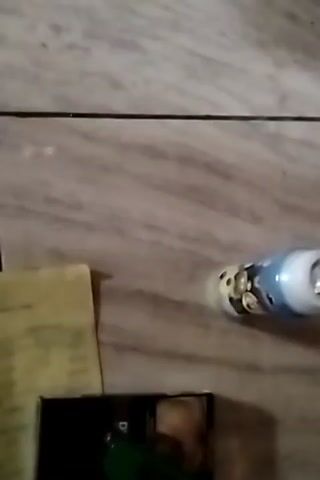A close-up, top-down view of a cluttered surface, likely a floor or table, with various objects scattered about. In the upper right quadrant, a cylindrical object with a light blue top and a white body, adorned with cartoon characters, is visible. To the left of this, a small, yellow piece of paper, possibly a receipt or note, is laid out. Below the yellow paper, a dark-colored rectangular object, possibly a pack of cigarettes or a small box, is present. A small portion of a green object is peeking out from beneath the dark object. The background consists of a marbled or tiled surface with beige and grey tones. The lighting is dim, casting shadows and obscuring some details. The camera angle suggests the shot was taken from directly above or at a slight downward angle. No people are visible in the frame. The scene appears to be indoors.