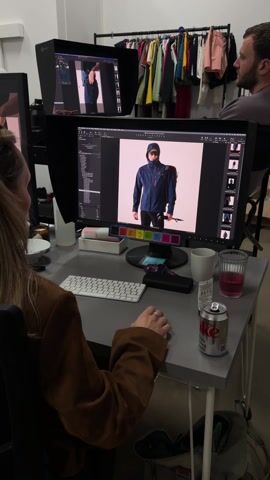 Here's a detailed description of the video:

The video shows a person sitting at a desk, editing a photo on a large computer monitor. The photo on the screen depicts a young man wearing a blue jacket and black pants against a white background. The editor has access to a control panel below the screen, with colored buttons. To the left, there is a white Apple keyboard and a collection of items including a small dish, a bottle of clear liquid, and a box. To the right, there is a white mug, a glass of pink liquid, a tube of some sort of cream, and a can of soda. 

There is a second computer monitor behind the main monitor. Behind both monitors, there is a clothing rack with various items of colorful clothing. A man is visible in the background standing near the clothing rack. The overall setting appears to be a studio or office environment.