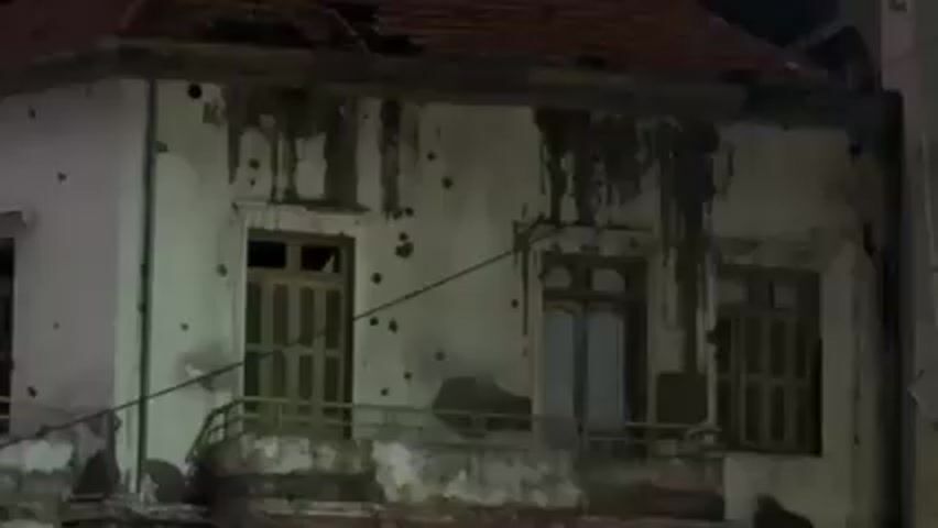 At night, the camera focuses on a two-story building with a partially damaged roof and multiple holes in its walls, potentially caused by bullets. The building’s windows are shuttered, and the paint is peeling. Dark stains streak down the walls. The camera then pans out to reveal the building is situated in an urban environment, surrounded by taller, more modern buildings with lit windows. A street with sparse traffic is visible, and a hand momentarily obscures the view. Streetlights illuminate the scene, enhancing the contrast between the old, damaged building and its modern surroundings.