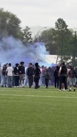 A group of young people stands on an artificial turf field, with a fence and trees in the background. Blue smoke is visible on the left side of the frame, near the fence and behind the crowd. Some individuals are casually walking, while others stand still. The sky is overcast.
