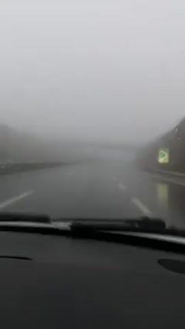 The video shows a view from inside a car, driving on a highway in very foggy conditions. Raindrops are visible on the windshield, which is being cleared by windshield wipers. The road is wet and lined with white stripes. An overpass is visible through the fog in the distance. There are green patches of vegetation on the sides of the road. The visibility is extremely poor.