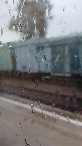 The video shows a close-up view of raindrops on a window. Through the water-streaked glass, a blurred scene of train tracks and a train can be seen. The train appears to be stationary or moving very slowly, and its colors are muted by the rain. The overall impression is one of a gray, damp day, possibly at a train station.
