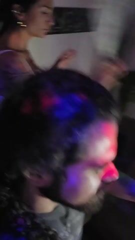 The video shows a person with long, curly hair from the shoulders up, focused on a woman in a white top dancing. She's in a dimly lit room with disco lights shining on her and the person recording. The room appears to be a living room with a couch and some artwork on the walls. The dancing woman moves her arms to the music, appearing energetic. The person filming is relatively still, holding the camera while observing the scene. There's a brief flash of another woman, also smiling, in the rotating disco lights. The mood is lively and celebratory.