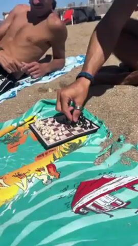 Two shirtless men are playing chess on the beach. A small travel chess set sits atop a colorful beach towel spread out on the sand. One man moves a black pawn on the board while the other watches. In the background, the beach stretches out under a sunny sky.