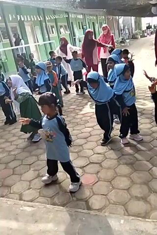 This is a vertical video. The first scene shows a group of young children in an outdoor courtyard, some wearing blue and white uniforms with headscarves. They are standing in rows and appear to be performing some kind of synchronized movements or dance. In the background, there is a green building and a white fence. The second scene shows a group of children gathered around a water fountain, with some appearing to be washing their hands. A woman in a red and white hijab is visible in the background, and other children are watching. The third scene focuses on a woman wearing a red hijab and a white top with red trim, performing a dance or some kind of movement in an outdoor covered area with tiled flooring. There is a speaker and some posters on the wall behind her. The final scene is a close-up of the water fountain. The video appears to be a compilation of different moments, possibly from a school event.