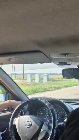 The video shows the interior of a car looking out through the windshield. A person's hands are visible on the steering wheel, which has a black cover. Outside the car, a light green train is passing by on railway tracks. The train appears to be moving from left to right in the frame. In the background, there are some white structures and green vegetation. The sky is visible and appears to be overcast.