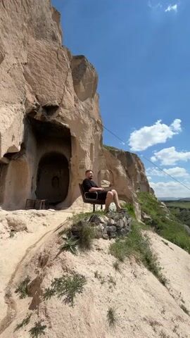 A man sits outdoors in a scenic, rocky landscape. He is seated on a dark chair placed on a small mound of rocks, overlooking a valley. Behind him are large, light-colored rock formations with carved cave-like openings, characteristic of the Cappadocia region. The sky is bright blue with scattered white clouds, indicating a sunny day. The man, wearing a black t-shirt and black shorts, has a beard and a short haircut. He looks relaxed and content, with a smile on his face as he gazes into the distance. Sparse green vegetation grows on the sandy ground around him. In the background, a white plastic container is visible near the rock structures.